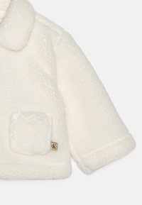 Valge kunstkarusnahast jakk, millel on kraega kaelus, üleliigselt suured varrukad, ruudukujuline tasku ja taskul väike brändi silt.