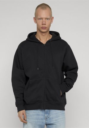 Jong persoon draagt een zwart zip-up hoodie met handen in de voorzakken en lichtblauwe spijkerbroek, staand tegen een effen achtergrond.