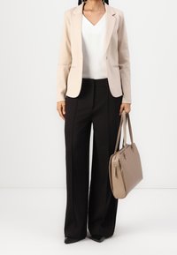 Blazer beige chiaro sopra una camicia bianca, abbinato a pantaloni larghi neri. Borsa beige con hardware argentato. Tessuti dalla superficie liscia.