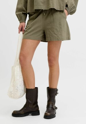 Persoon draagt olijfgroene shorts en bijpassende top, houdt een witte kanten tas vast en draagt zwarte leren laarzen tot halverwege de kuit met gespen.