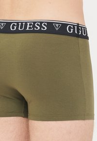 Maslinaste zelene bokserice napravljene od glatkog materijala, s crnim pojasom s bijelim "GUESS" logotipom i kontrastnim obrubom na vrhu.