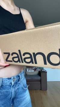 Hnedá lepenková krabica s čiernym brandovaním "zalando", držaná osobou. V pozadí je pohovka a drevená podlaha.