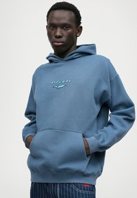 Felpa blu realizzata in morbido tessuto, con una tasca frontale e cappuccio regolabile con cordino. Sul petto è ricamato un logo in colori chiari.