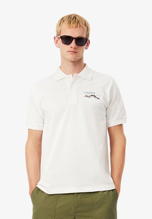 Jeune homme portant des lunettes de soleil noires, un polo Lacoste blanc avec un logo de montagne et un pantalon vert olive, se tenant avec les mains dans les poches.