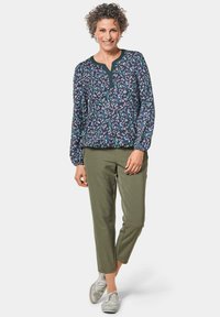 Bluse mit floralem Druck in Marineblau mit grünen und weißen Akzenten, verfügt über einen V-Ausschnitt und lange, geraffte Ärmel. Kombiniert mit olivgrünen Hosen.