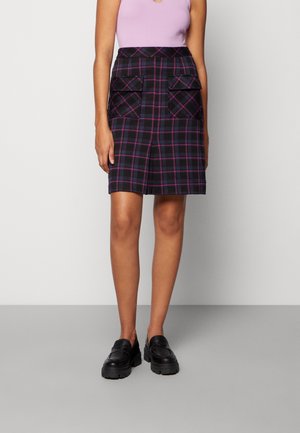 Femme portant une jupe à carreaux noire et violette avec des poches à rabat sur le devant, un haut sans manches lilas, et des mocassins noirs épais.