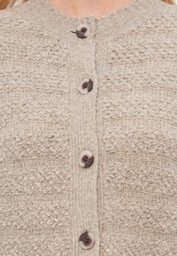Lys beige cardigan med teksturert stoff, med rund hals og fem brune knapper med lysere knappdeksler.