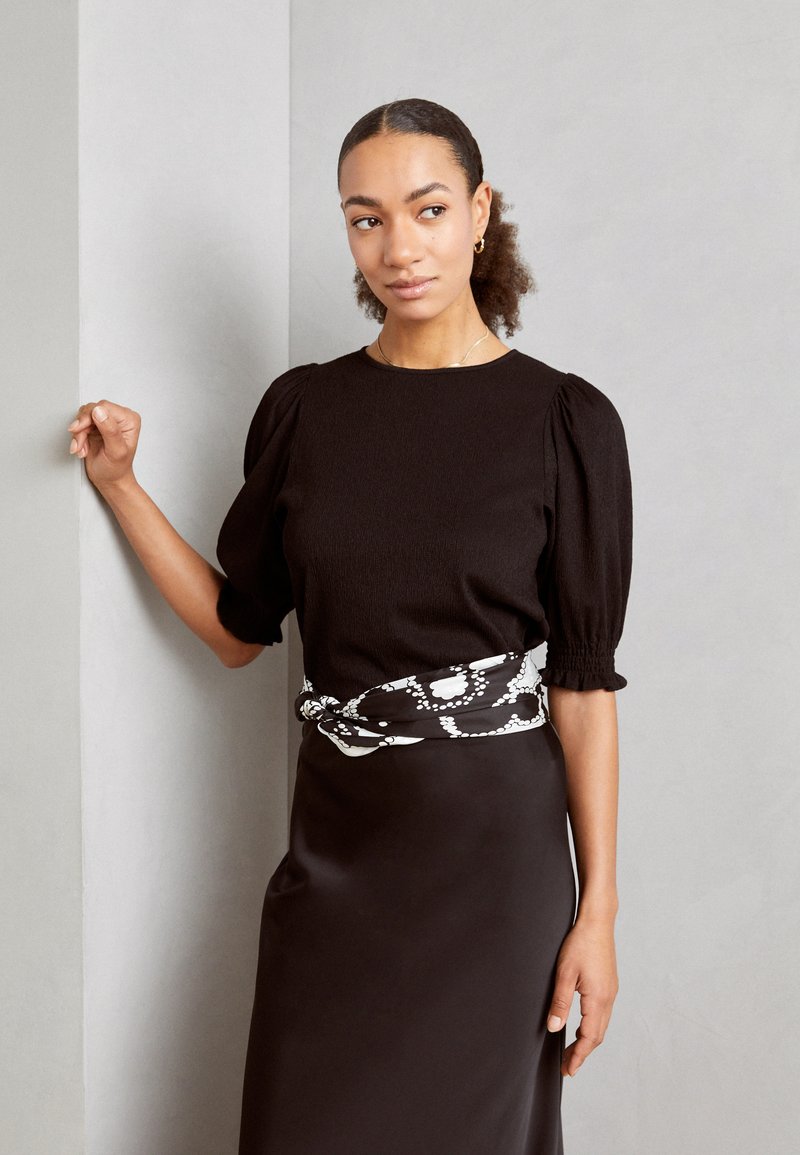 Black textured top with puff sleeves, paired with a sleek brown skirt. White patterned belt adds contrast. Model poses against a wall.