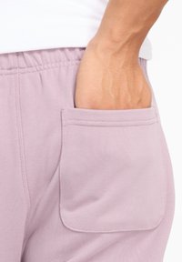 Hellviolette Jogginghosen mit elastischem Bund, seitlichen Taschen an den Nähten und einem weichen, strukturierten Stoff. Sichtbare Hand in der Tasche.