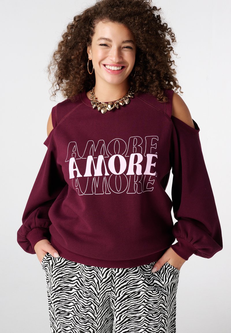Sweat-shirt bordeaux avec épaules découpées et inscription "AMORE" en différentes polices. Associé à un pantalon imprimé zèbre noir et blanc, avec poches.