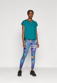 Κοντομάνικο τοπ σε πράσινο-μπλε απόχρωση, συνδυασμένο με πολύχρωμους κολάν με λουλούδια σε μπλε και μωβ σχέδιο, διακοσμημένα με ανακλαστικές ρίγες. Μαύρα αθλητικά παπούτσια.