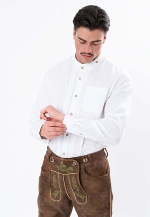 Man wearing white buttoned shirt and traditional brown leather lederhosen adjusts cuff with downward gaze on white background.