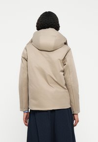 Beige, párnázott dzseki nagy, struktúrált kapucnival, sima textúrával és patentos záróval az ujjakon, hátulról nézve.