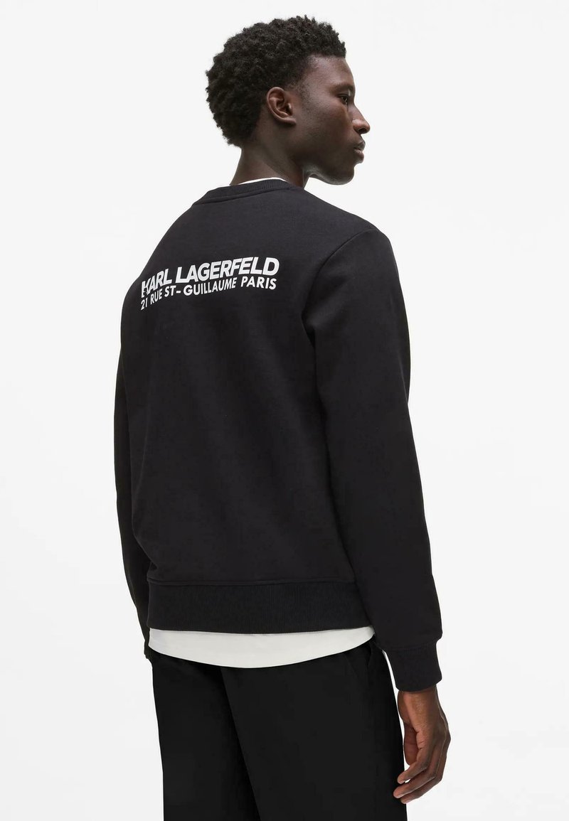 Jeune homme vu de profil portant un sweat-shirt noir avec l'inscription "Karl Lagerfeld 21 Rue St-Guillaume Paris" imprimée dans le dos, chemise blanche en dessous.