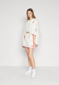 Kuttet hvit jakke med oransje detaljer, kombinert med en hvit crop-top og et hvitt skjørt med oransje striper. Hvite sneakers fullfører antrekket.