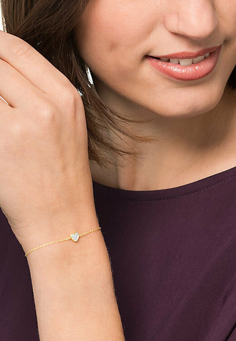 Goldene Kettenarmband mit einem kleinen Herzanhänger, der eine strukturierte Oberfläche und einen funkelnden Akzent in der Mitte aufweist, am Handgelenk getragen.