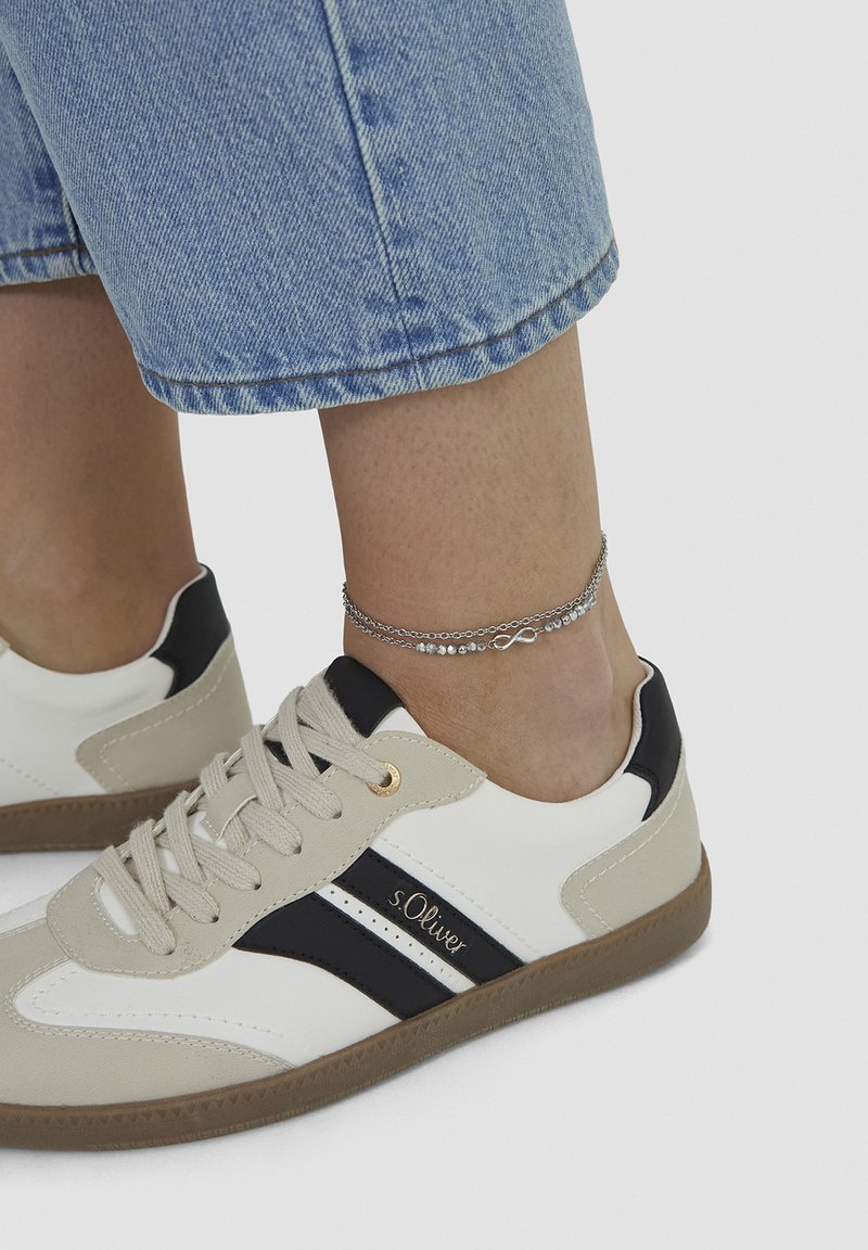 Ben iført beige og hvide s.Oliver sneakers med sorte striber, kombineret med denim bukser og sølvfarvet uendelighedskæde om anklen.
