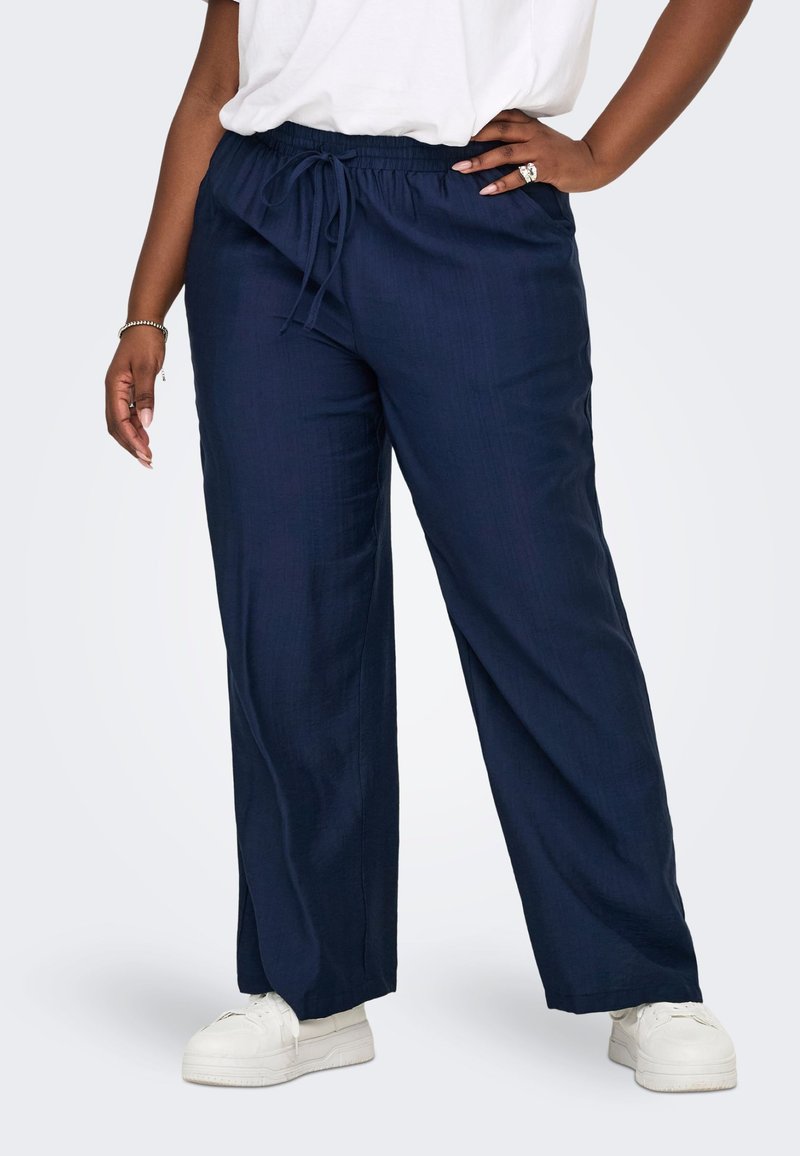 Άτομο που φοράει σκούρο μπλε παντελόνια με φαρδιά μπάτζα και κορδόνι, λευκά αθλητικά παπούτσια και λευκό τοπ, στέκεται με το ένα χέρι στη μέση.