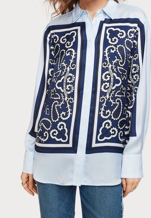 Jasnoniebieska bluzka z długim rękawem z ozdobnym wzorem paisley w kolorze granatowym i białym oraz napisem, noszona nie włożona do środka, z niebieskimi dżinsami.