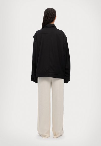 Black oversized jacket with a collared neckline and dropped shoulders, paired with cream wide-leg trousers, seen from the back.