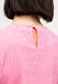 Πίσω μέρος του λαιμού και κοντά σκούρα μαλλιά πάνω από μια ροζ μπλούζα με υφή, με μια μικρή άνοιγμα σε σχήμα κλειδαρότρυπας που κλείνει με κουμπί καλυμμένο με ύφασμα.