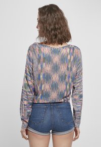 Donna con capelli castani mossi di media lunghezza, che indossa un maglione a maglia multicolore e shorts di jeans arrotolati, di spalle contro uno sfondo semplice.