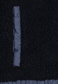 Svart teksturert fleece-stoff med panel i blå nylon; har et vertikalt spalteutskjæring i fleece, som fremhever kontrasten mellom materialene.