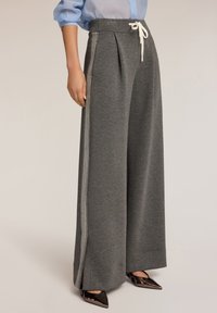 Grijze wijde sweatpants van zacht materiaal met een geplooid ontwerp, voorzien van een witte trekkoord en zijstrepen. Zwarte puntige schoenen zichtbaar.