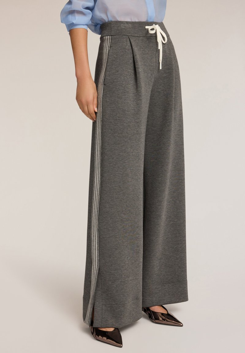 Grijze wijde sweatpants van zacht materiaal met een geplooid ontwerp, voorzien van een witte trekkoord en zijstrepen. Zwarte puntige schoenen zichtbaar.