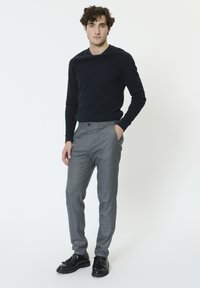 Fiatal férfi fekete pulóverben, szürke nadrágban és fekete bojtos mokaszinokban, egyik kezével a zsebében áll egy sima fehér háttér előtt.