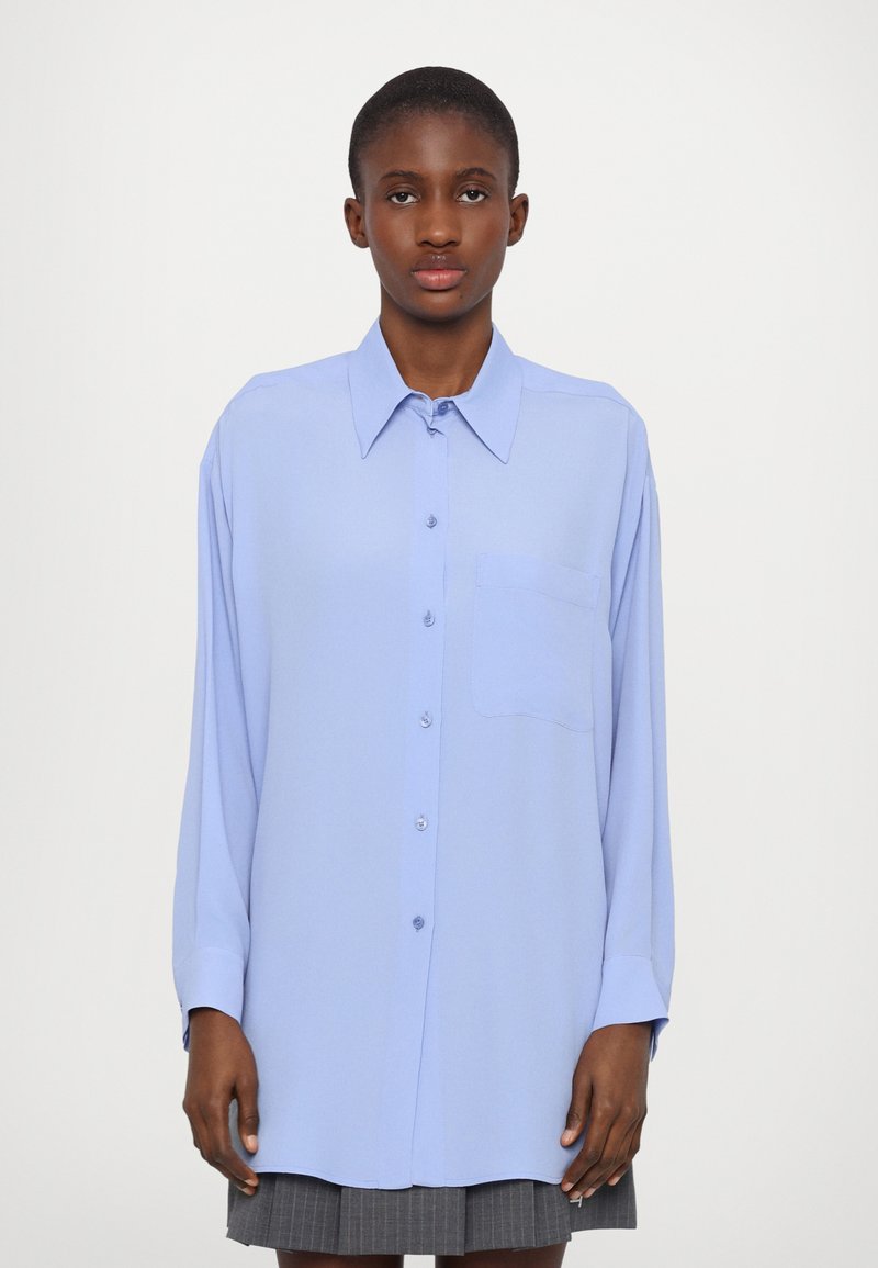 Camicia azzurra a maniche lunghe realizzata in tessuto liscio, con colletto a punta, chiusura con bottoni e una singola tasca sul petto.