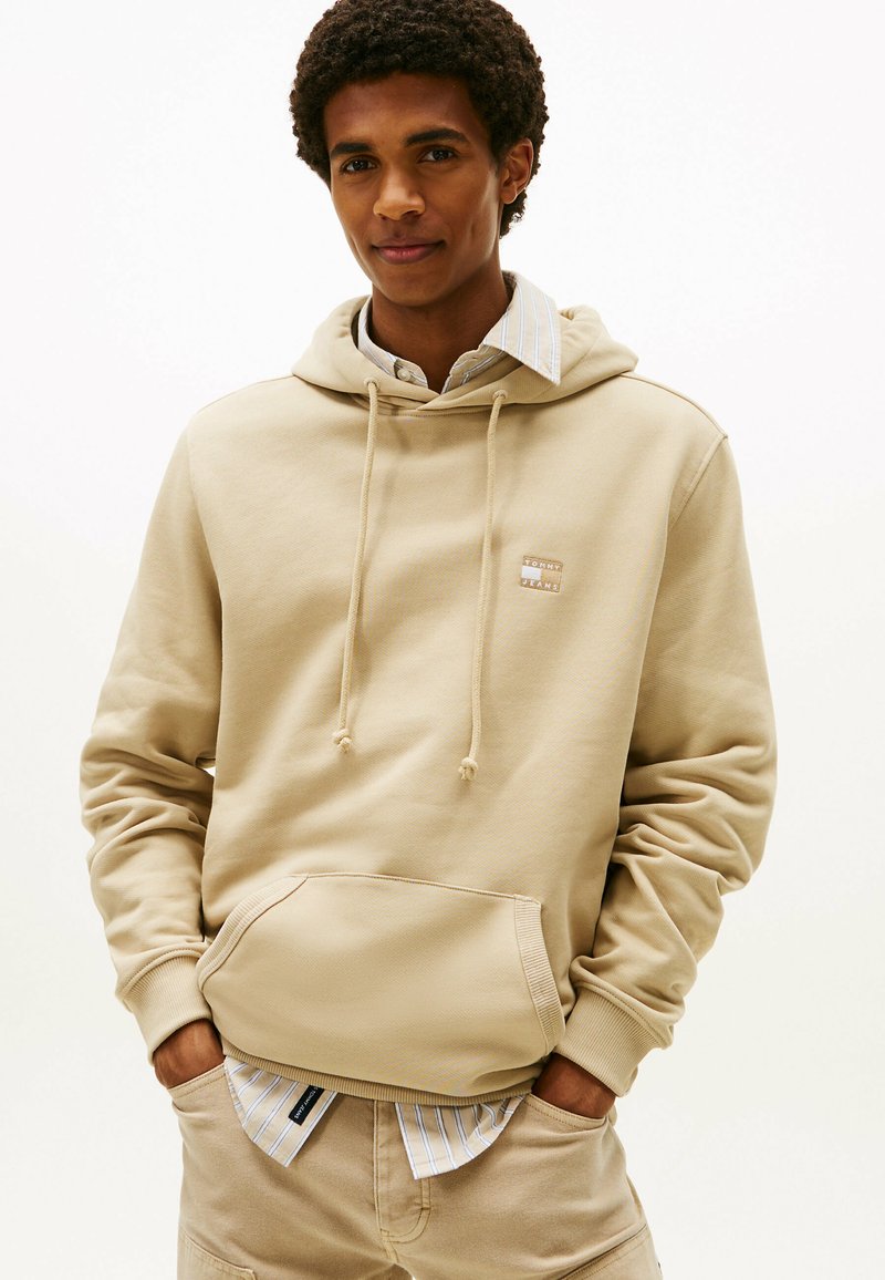 Beige Baumwoll-Hoodie mit Kordelzügen, Fronttasche und Logo-Patch. Wird über einem hellblauen und weißen gestreiften Shirt getragen. Glatte Textur.