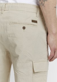 Pantalons cargo beiges en tissu léger, avec une poche arrière, une fermeture à bouton et une étiquette INDICODE marron sur la taille.