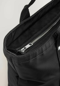 Schwarze Stofftasche mit einem Reißverschluss oben, strukturierten Griffen und einer Innenauskleidung, die ein silbernes Reißverschlussdetail zeigt.
