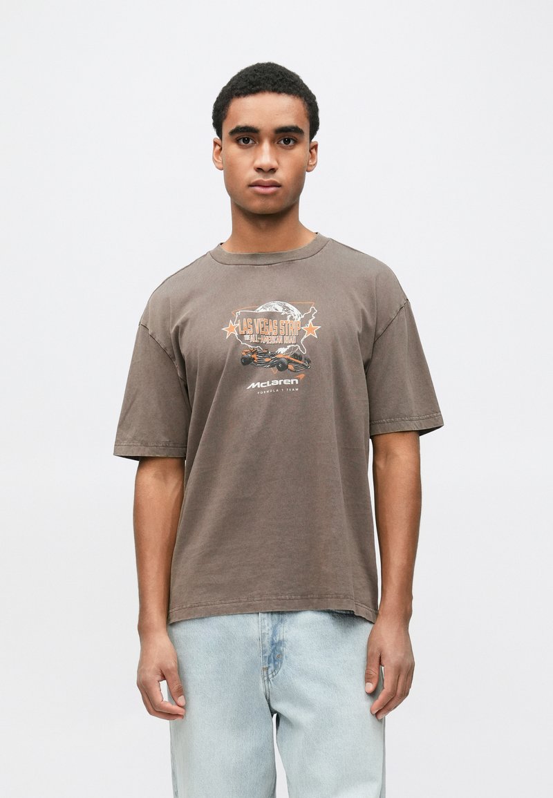 Καφέ βαμβακερό t-shirt με στρογγυλή λαιμόκοψη, κοντά μανίκια και γραφικό σχέδιο με μια υδρόγειο σφαίρα και κείμενο, σε συνδυασμό με ανοιχτό μπλε τζιν παντελόνι.