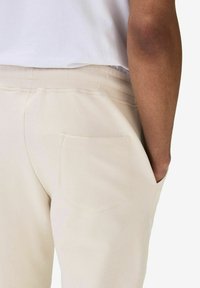 Beige sweatpants med en strukturerad yta, med en bakficka och ett elastiskt midjeband. Bäraren har en hand i fickan.