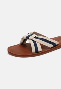 Sandales plates avec une base en cuir marron et des brides tissées beige avec des rayures bleu marine. Les brides se croisent sur le pied, assurant un maintien sûr.