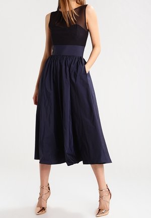 Frau trägt ein ärmelloses schwarz-marineblaues Midi-Kleid mit transparentem Oberteil und goldenen Riemchen-Hochschuhen, steht mit einer Hand in der Tasche.