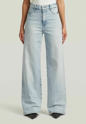 Personne portant un jean bleu clair taille haute à jambes larges avec des chaussures noires pointues, debout devant un fond clair uni.