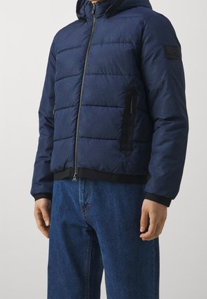 Mand iført navyblå quiltet jakke med hætte, sort lynlås og lommedetaljer, kombineret med blå jeans, mod en ensfarvet baggrund.