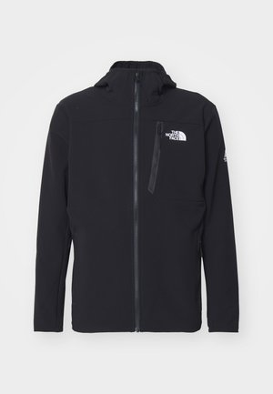 Casaco softshell preto com fecho à frente, colarinho alto e bolso no peito; tem um logotipo no lado esquerdo e tecido texturizado.