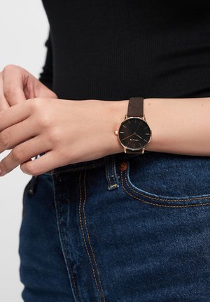 Schwarze Uhr mit rundem Zifferblatt, goldenen Akzenten und braunem Lederband, am Handgelenk getragen, kombiniert mit blauen Jeans.