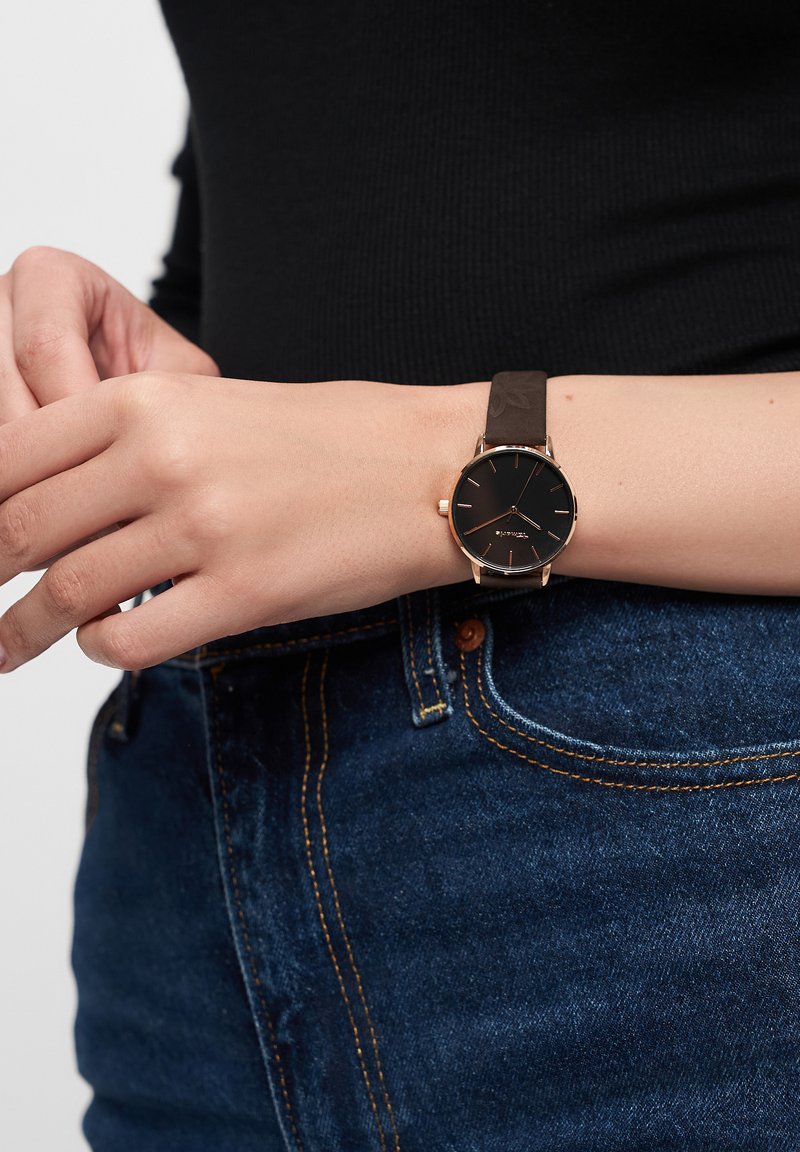 Montre noire avec un cadran rond, accents dorés, et bracelet en cuir marron, portée au poignet, associée à un jean en denim bleu.