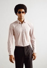 Světle růžová košile na knoflíky s diagonálními bílými pruhy, spárovaná s černými kalhotami. Model nosí tmavé sluneční brýle a má afro účes.