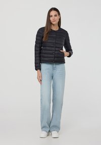 Giacca imbottita nera con collo rotondo e chiusura a bottoni, caratterizzata da una texture trapuntata orizzontale, indossata sopra jeans dritti blu chiaro.