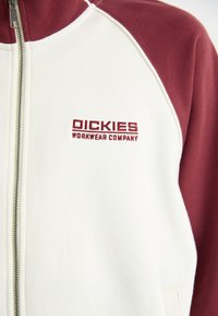 Maroon and white zip-up sweatshirt with ribbed collar, featuring embroidered "Dickies Workwear Company" logo in red on the chest. Soft texture.