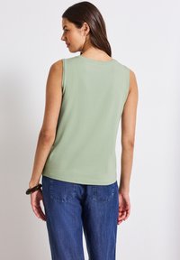 Top senza maniche verde chiaro con scollatura rotonda, texture liscia e orlo a strati; abbinato a pantaloni in denim blu con tasche posteriori.