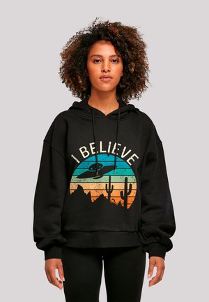Black hoodie featuring a colorful circular graphic with mountains, cacti, and the phrase "I BELIEVE" in a retro font. Soft fabric texture.
