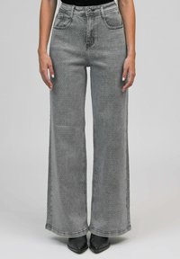 Femme portant un jean gris taille haute et large avec de petites embellissements en pois, associé à des chaussures noires à bout pointu et un top noir.