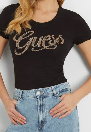 Femme portant un t-shirt noir ajusté avec le logo "Guess" en strass dorés, associé à un jean taille haute bleu clair, les mains dans les poches.